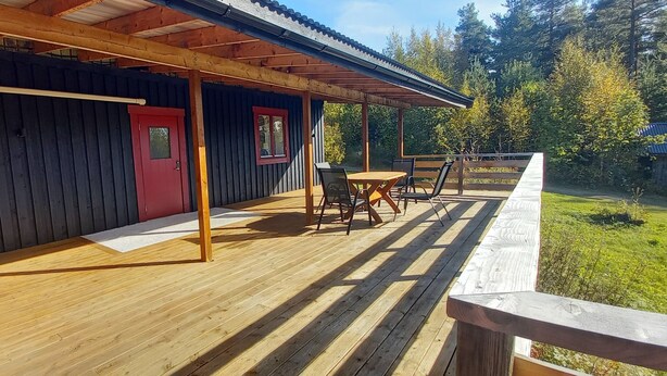 Um deck de madeira com área de estar coberta, uma porta vermelha e um ventilador à esquerda.