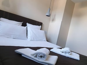 Un lit bien fait, avec des draps blancs et deux serviettes pliées posées sur une couverture de couleur foncée.