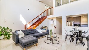 Een moderne woonkamer met een grijze modulaire bank, een ronde salontafel en een eethoek met een glazen tafel en metalen stoelen. Een houten trap leidt naar een hoger gelegen verdieping.