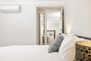 A bedroom with a bed, a bedside lamp, and a view of a hallway with a door and a chair.