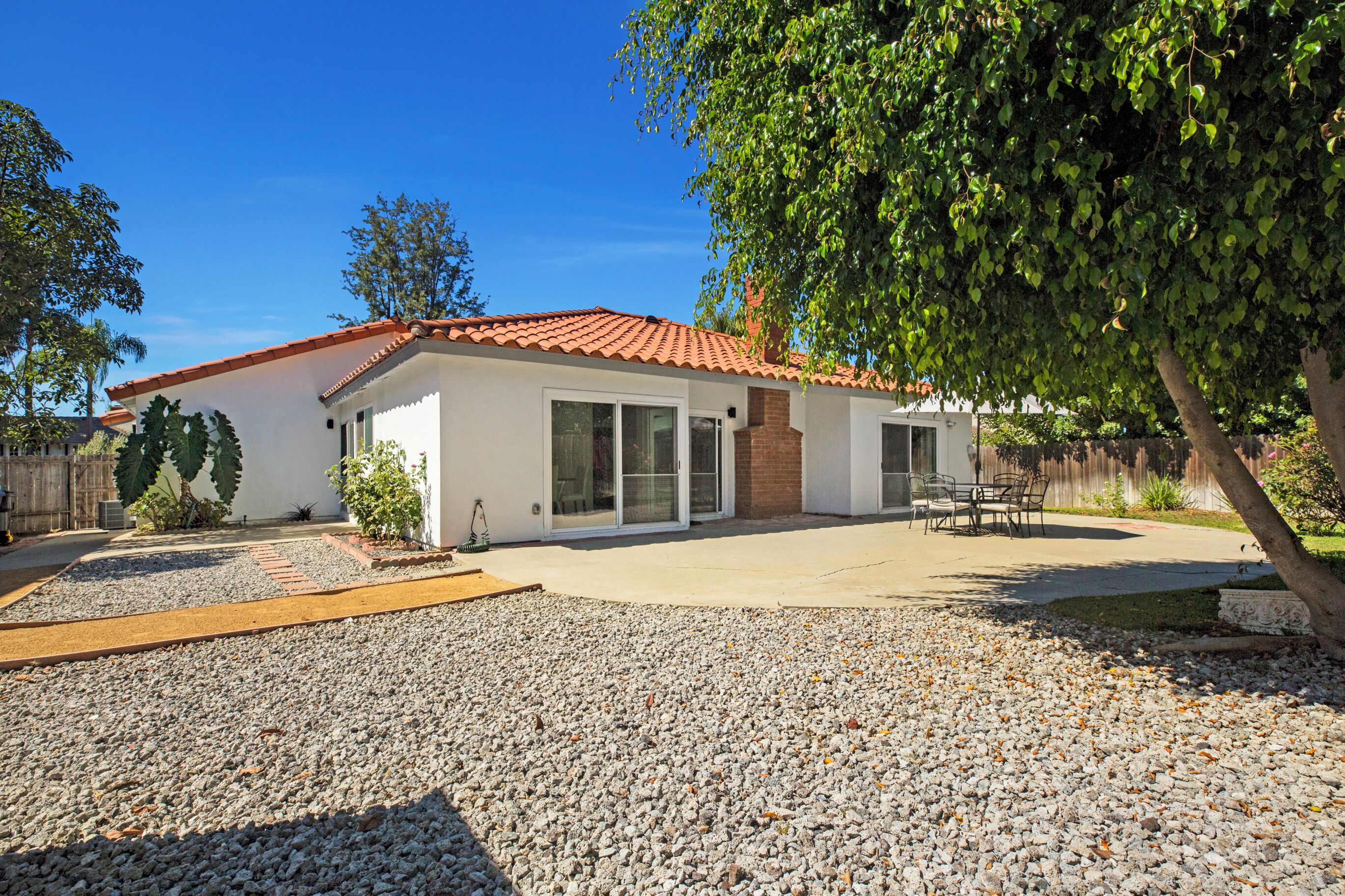 Una casa de un piso con techo de tejas rojas, un patio con mobiliario exterior y un camino de acceso de grava.