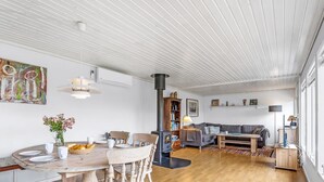 Amplia sala de estar con una mesa de comedor de madera, sillas y un sofá. Se ve una chimenea al fondo, y hay estanterías y objetos decorativos en las paredes.
