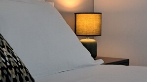 A neatly made bed with white linens and a patterned pillow, a bedside lamp with a textured shade, and a dark nightstand. - Cosy bilo close to the centre (Urban 9) (Lugano)