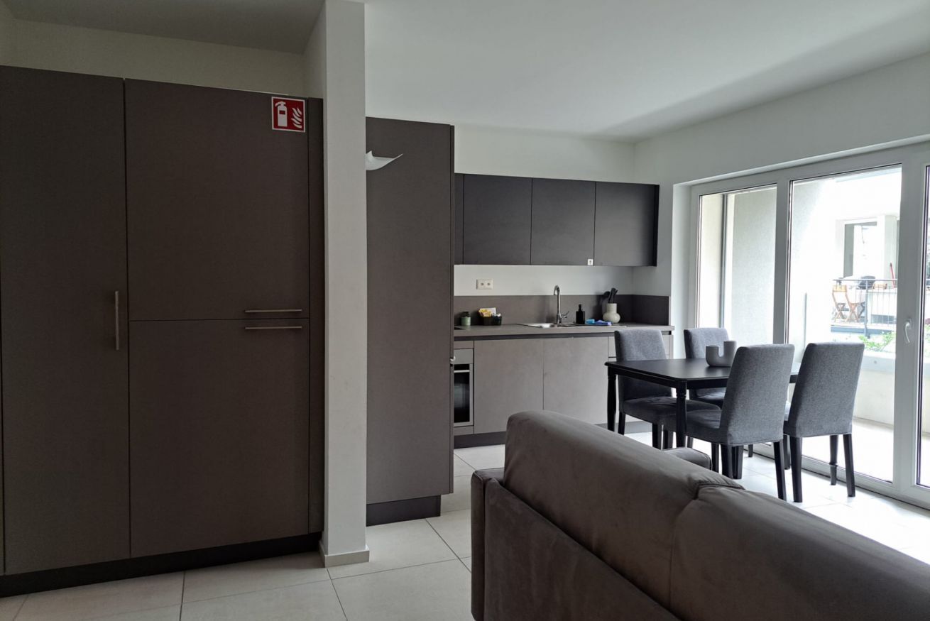 Een moderne keuken met donkere kastjes, een eethoek met een tafel en stoelen, en een bank in een lichte kamer met grote ramen.