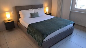 Una cama bien tendida con cabecera gris y ropa de cama blanca, dos lámparas de noche y una ventana con persianas.