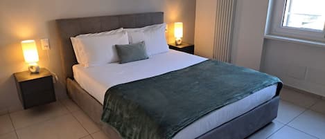 A neatly made bed with a grey headboard and white bedding, two bedside lamps, and a window with blinds.