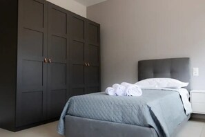 Een slaapkamer met een grote donkere garderobe, een bed met een grijs geborduurd sprei, en netjes opgerolde witte handdoeken op het bed.