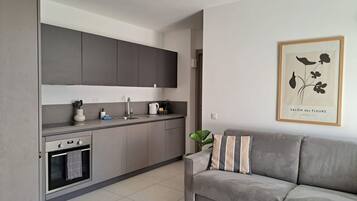 A modern kitchen with grey cabinets, a built-in oven, and a sink. There is a grey sofa with a cushion and a framed picture on the wall.