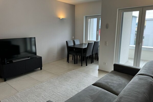 A modern living room with a flat-screen TV, a dining table with chairs, a sofa, and a sliding glass door leading to a balcony.