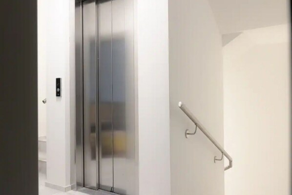A modern elevator with a metallic finish, a handrail on the right, and a control panel on the left.