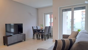 A modern living room with a flat-screen TV, a sofa, a dining table with chairs, and a large window with a view of buildings. - Cosy bilo close to the centre (Urban 5) (Lugano)