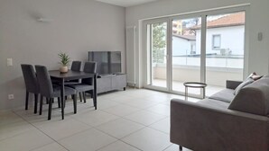 A modern living room with a dining area, a sofa, a TV, and a sliding glass door leading to a balcony. - Two-room apartment in new building in Lugano (Urban Residence 6) (Lugano)