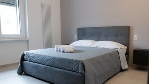 A neatly made bed with a grey duvet and white pillows, a radiator on the wall, and a window with blinds. - Two-room apartment in new building in Lugano (Urban Residence 6) (Lugano)
