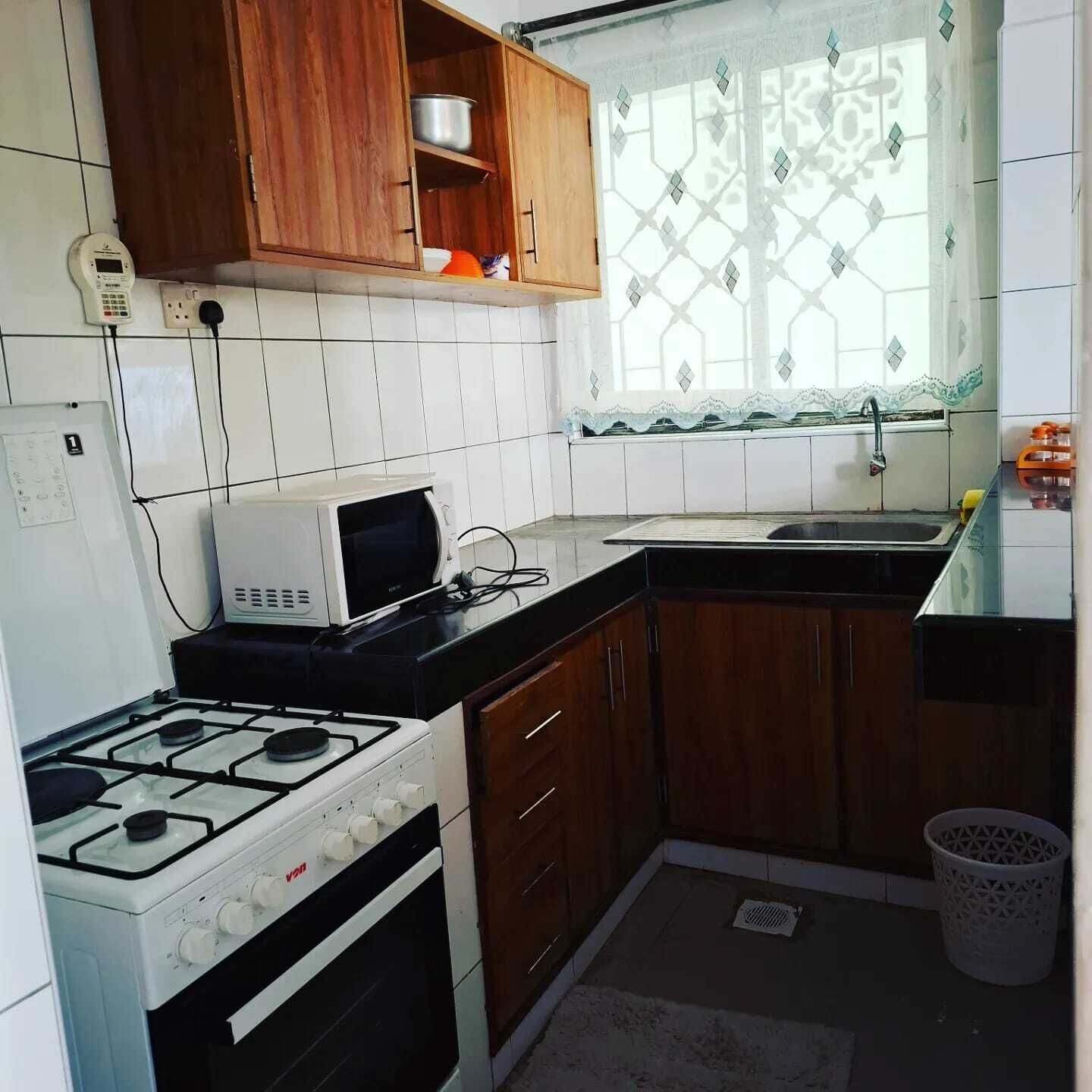 Een compacte keuken met een gasfornuis, een magnetron, houten keukenkastjes en een raam met gordijnen met een patroon.