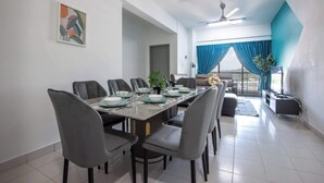 A modern dining area with a long table set for four, surrounded by grey chairs. There is a sofa and a television in the background. - Modern 3BR Near Danga Bay JB Free Parking (Johor Bahru)