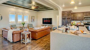 A spacious living room with a brown leather sofa, a coffee table, and a TV displaying 'NETFLIX'. There's a kitchen area with wooden cabinets and a dining table.