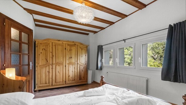 A bedroom with a wooden wardrobe, a bed with white linens, a wooden ceiling with exposed beams, and a window with a view of greenery. - Pretty chalet-style studio (Euseigne)