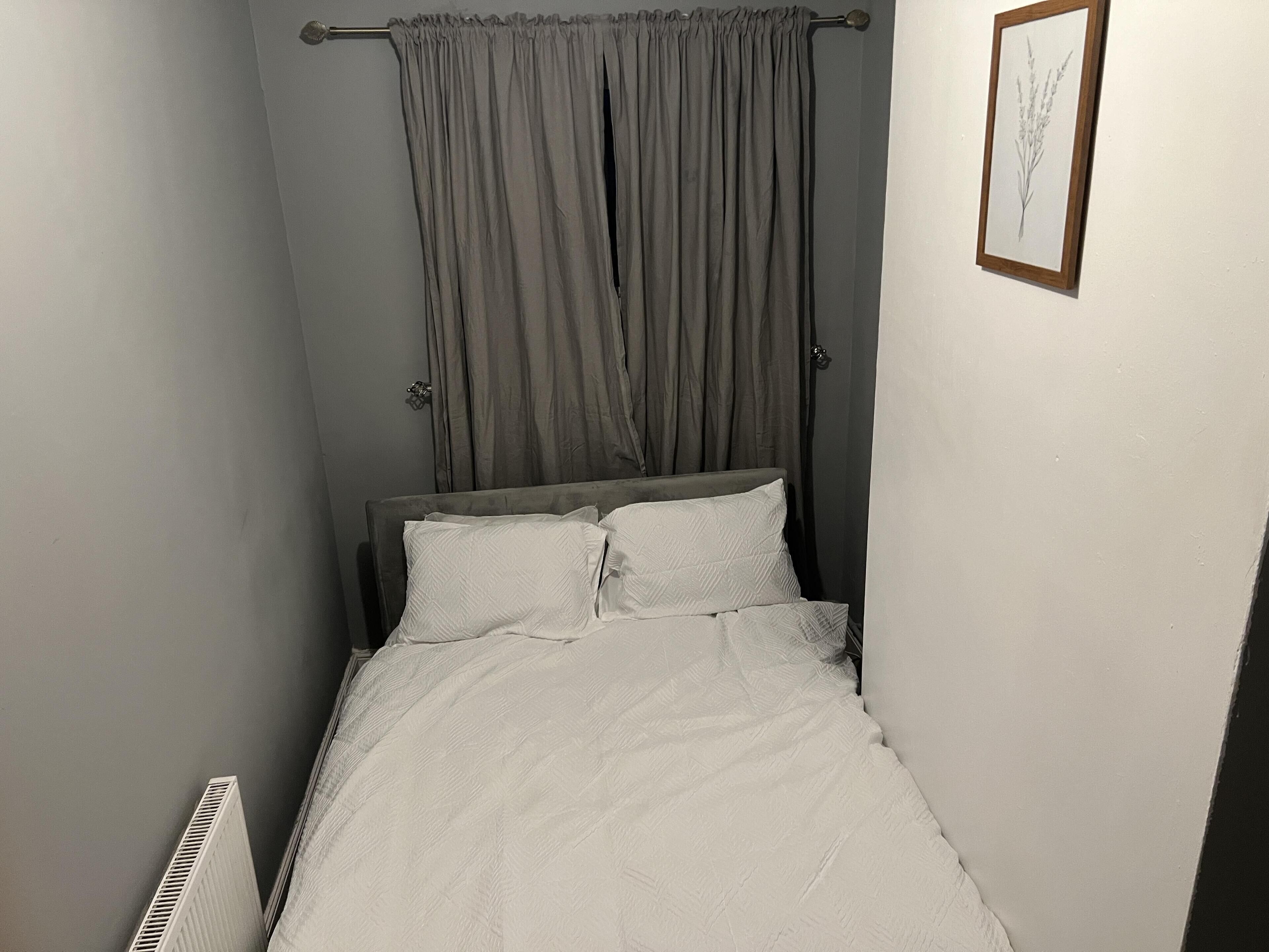 A narrow room with a bed, grey curtains, and a framed botanical print on the wall.