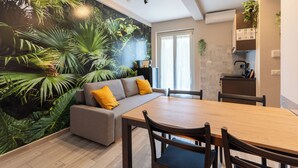 Ein modernes Wohnzimmer mit einer großen Wandmalerei von tropischen Pflanzen, einem grauen Sofa mit gelben Kissen, einem Holzesstisch und passenden Stühlen.