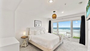 A bedroom with a large bed, a bedside table, a lamp, a framed picture, and a view of the beach through large windows. - Ocean Front Oasis - Team Joseph Ellen (Jacksonville Beach)