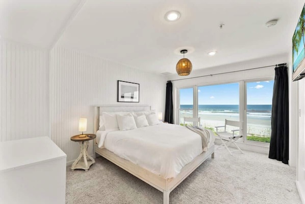 A bedroom with a large bed, a bedside table, a lamp, a framed picture, and a view of the beach through large windows.