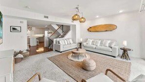 A spacious living area with a white sofa, a round coffee table, and a wooden surfboard on the wall. - Ocean Front Oasis - Team Joseph Ellen (Jacksonville Beach)