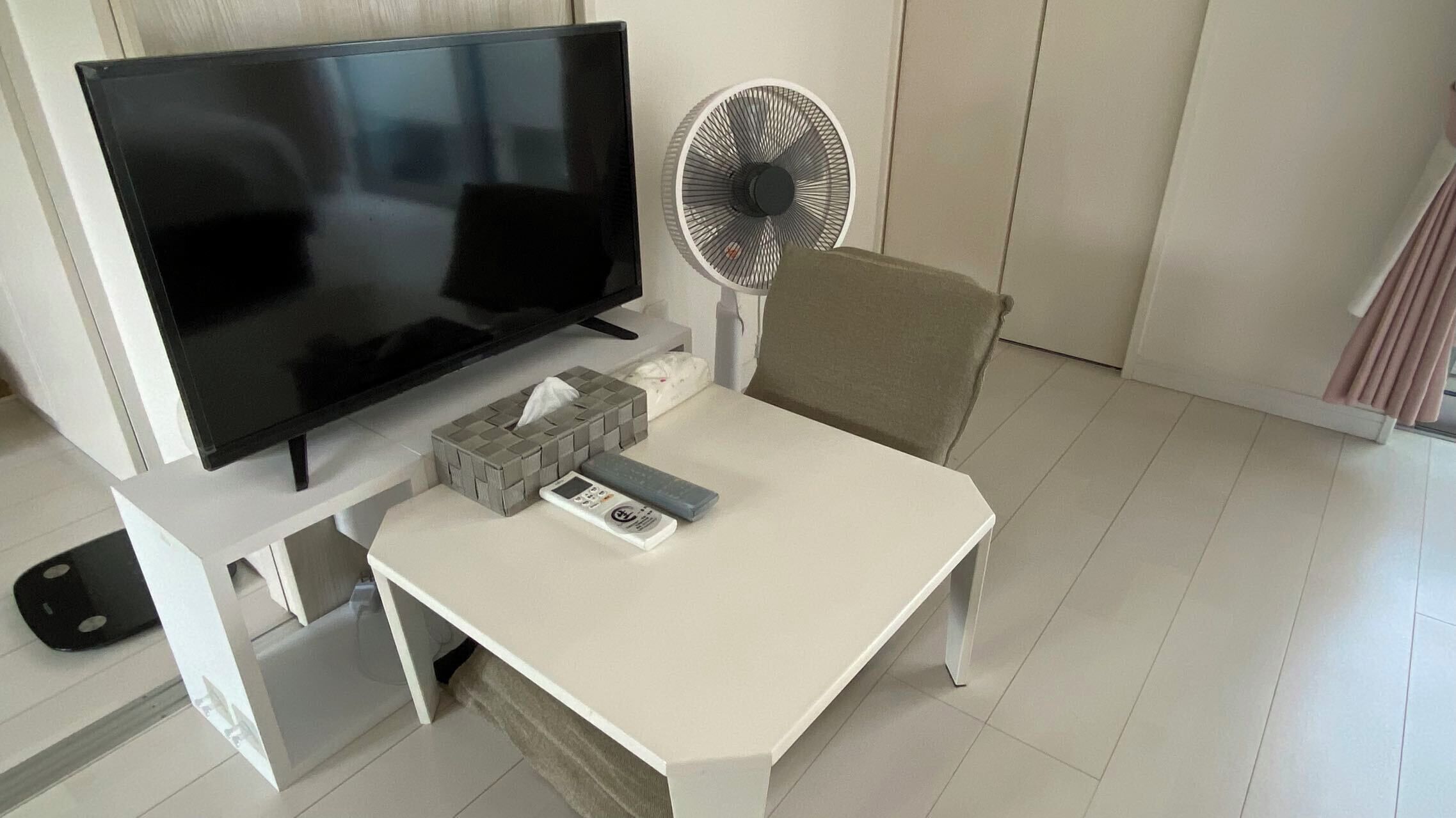 Een kamer met een flatscreen televisie, een witte tafel, een afstandsbediening, een zakdoekendoos en een ventilator.