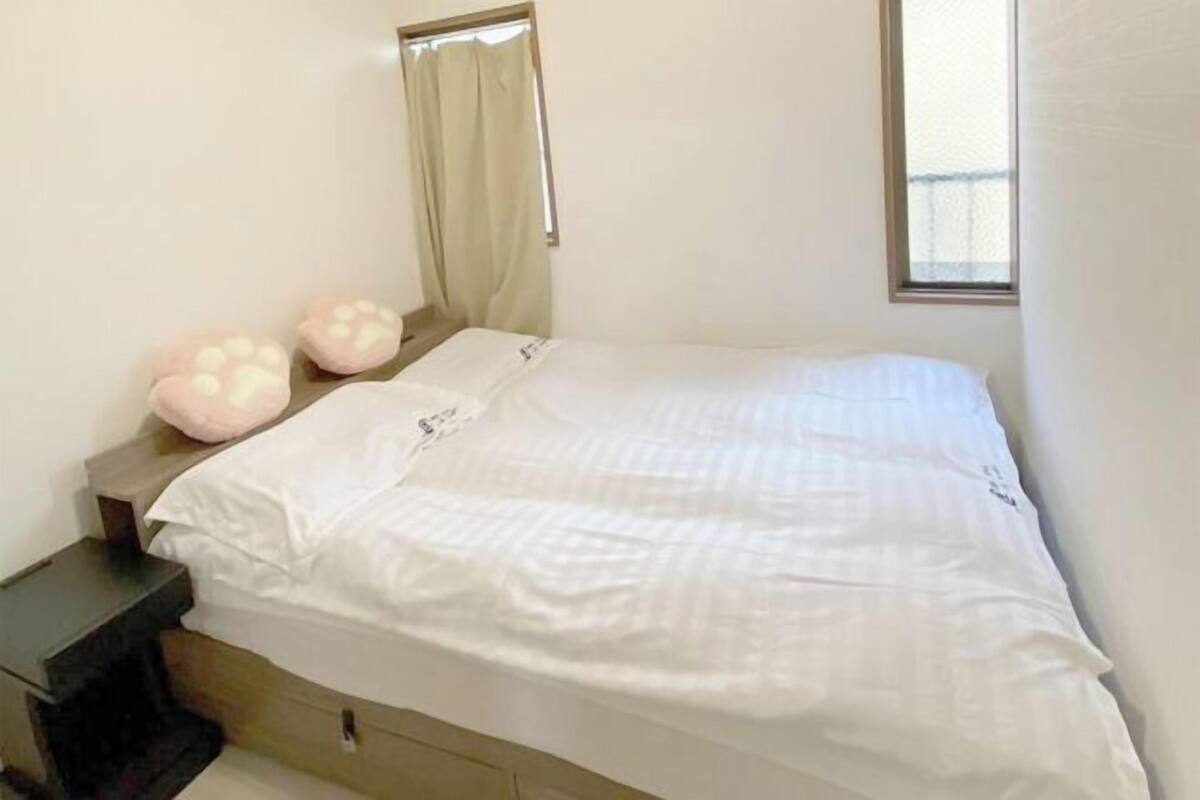 A single bed with a white mattress and pillows, a small bedside table, and a window with a curtain.