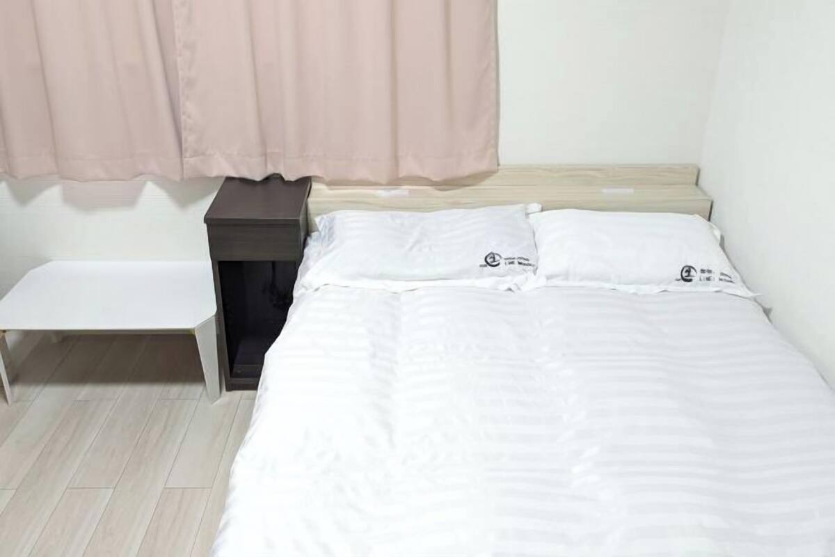 A single bed with white bedding, a bedside table, a small white bench, and a pink curtain.