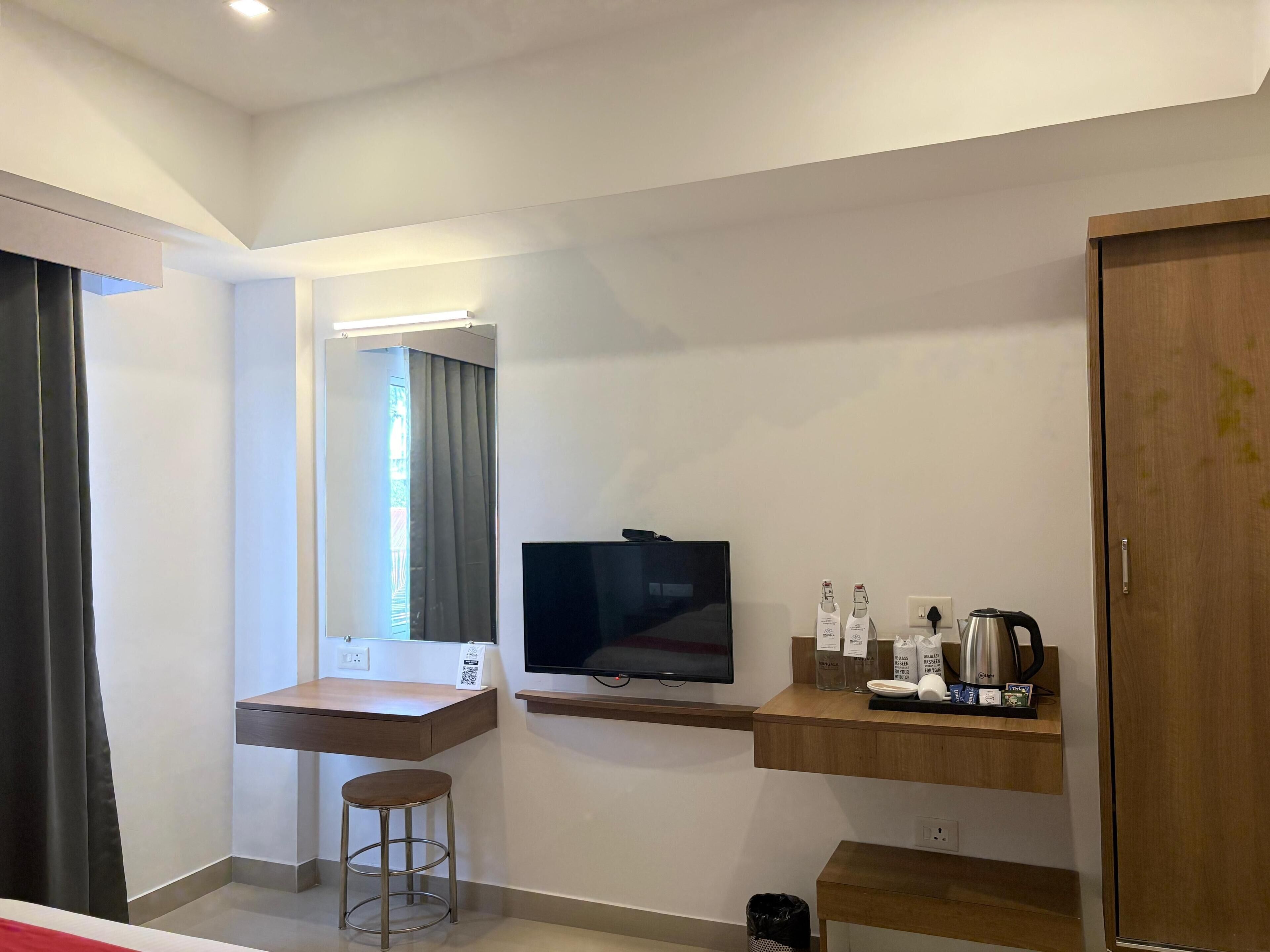 A modern hotel room with a flat-screen TV, a wooden desk, a stool, a small shelf with toiletries, and a wardrobe.