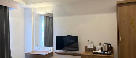 A modern hotel room with a flat-screen TV, a wooden desk, a stool, a small shelf with toiletries, and a wardrobe.