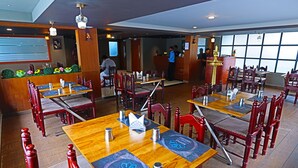 A restaurant with wooden tables and red chairs, a reception desk, and a large window in the background. - HOTEL MANGALA TEMPLE VIEW (Chavakkad)