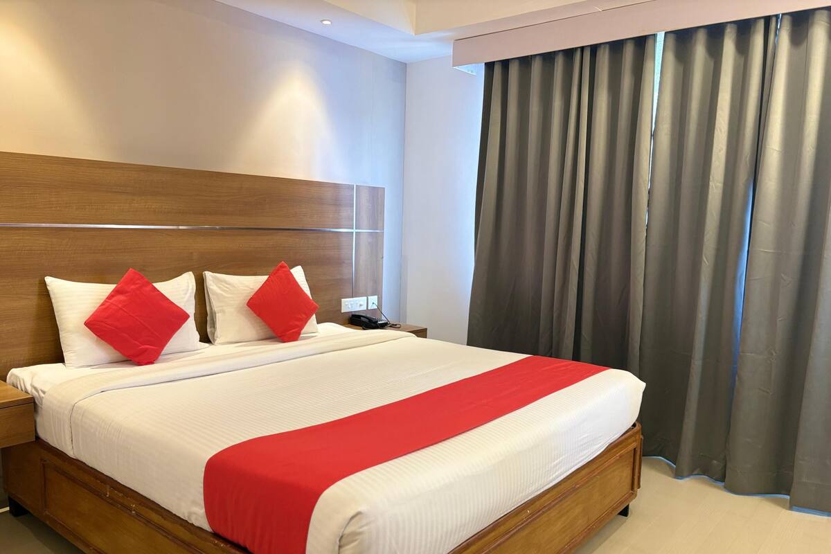 Habitación de hotel con cabecera de madera, cama grande con ropa blanca y detalles rojos, mesita de noche con teléfono y vista a cortinas grises.