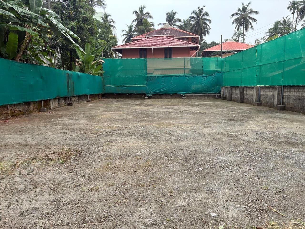 A construction site with a red-roofed building, green netting, and surrounding vegetation.