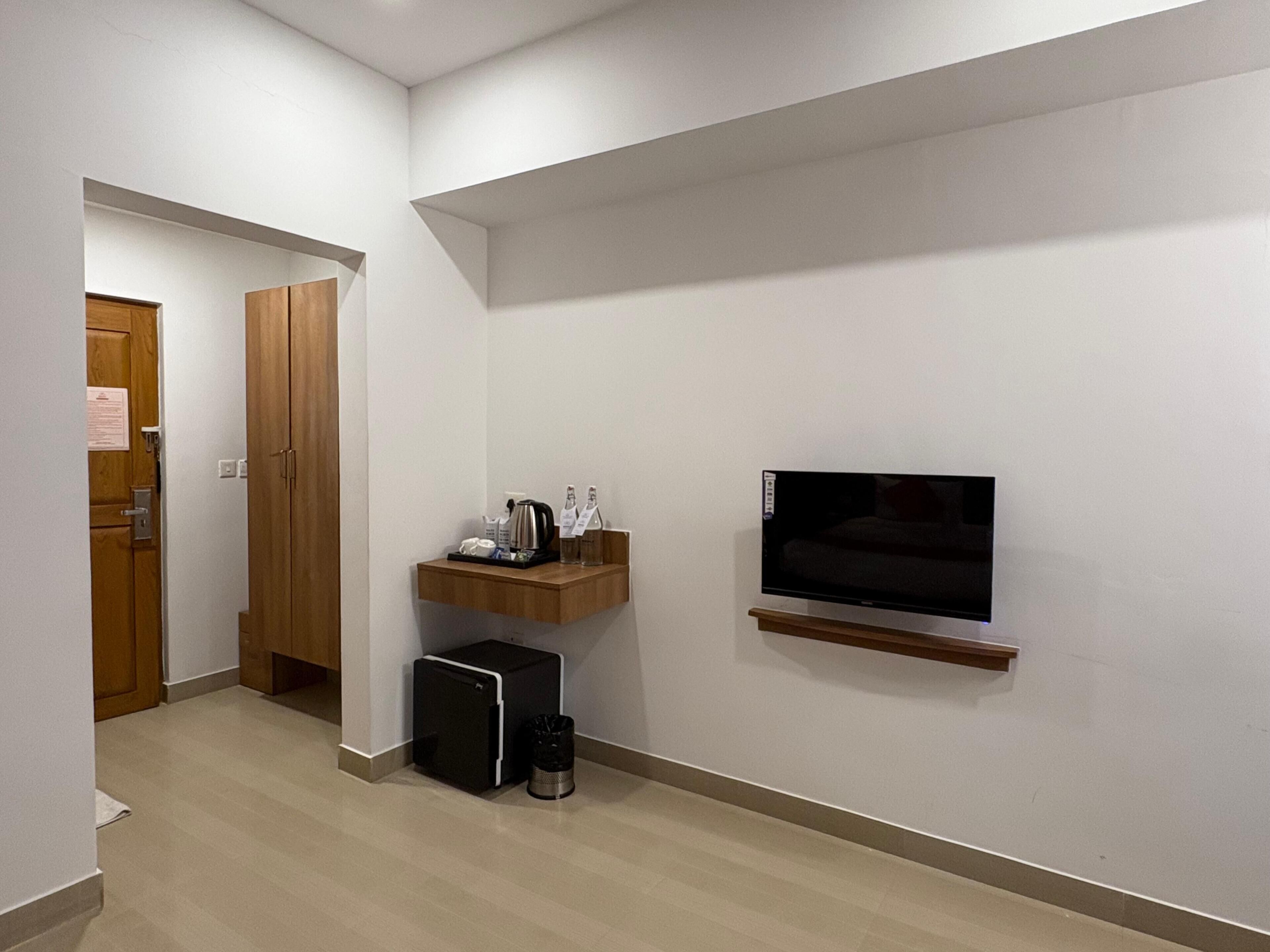 A hotel room with a wooden door, a small kitchenette, a TV mounted on the wall, and a wooden shelf.