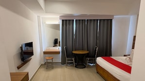 A hotel room with a bed, a TV mounted on the wall, a small table with two chairs, a desk with a chair, and a curtain. - HOTEL MANGALA TEMPLE VIEW (Chavakkad)