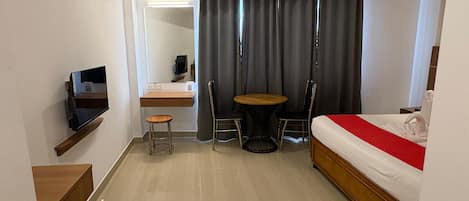 Hotellihuone, jossa on sänky, seinälle asennettu televisio, pieni pöytä ja kaksi tuolia, työpöytä ja tuoli sekä verho.