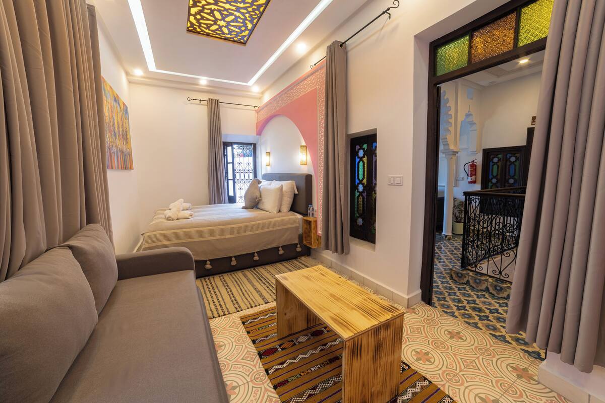 Une chambre d’hôtel moderne avec un canapé, un lit avec des oreillers, une table de chevet en bois, des tapis à motifs et un luminaire décoratif au plafond.