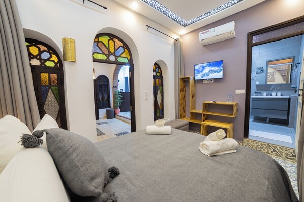 A hotel room with a large bed, a TV, a wooden shelf, and a bathroom visible through an arched doorway. - Iguana Azul (Chefchaouen)
