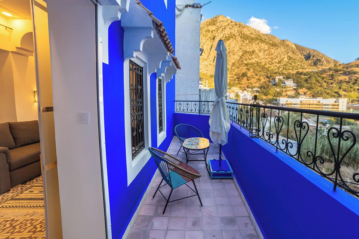 Un balcon aux murs bleus, avec une chaise, une petite table et une vue sur un paysage montagneux.