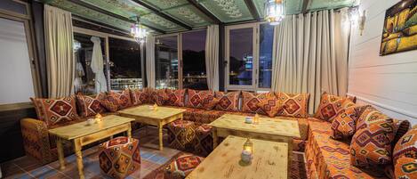 A cozy seating area with patterned cushions and wooden tables, large windows with curtains, and a city view at night.
