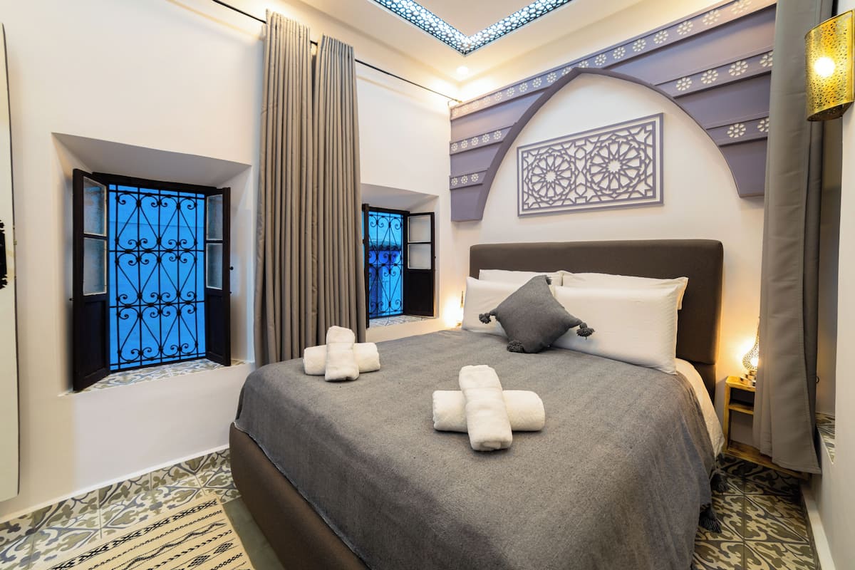 Une chambre d’hôtel avec un grand lit, deux fenêtres ornées de grilles décoratives, un sol à motifs et un élément décoratif mural.