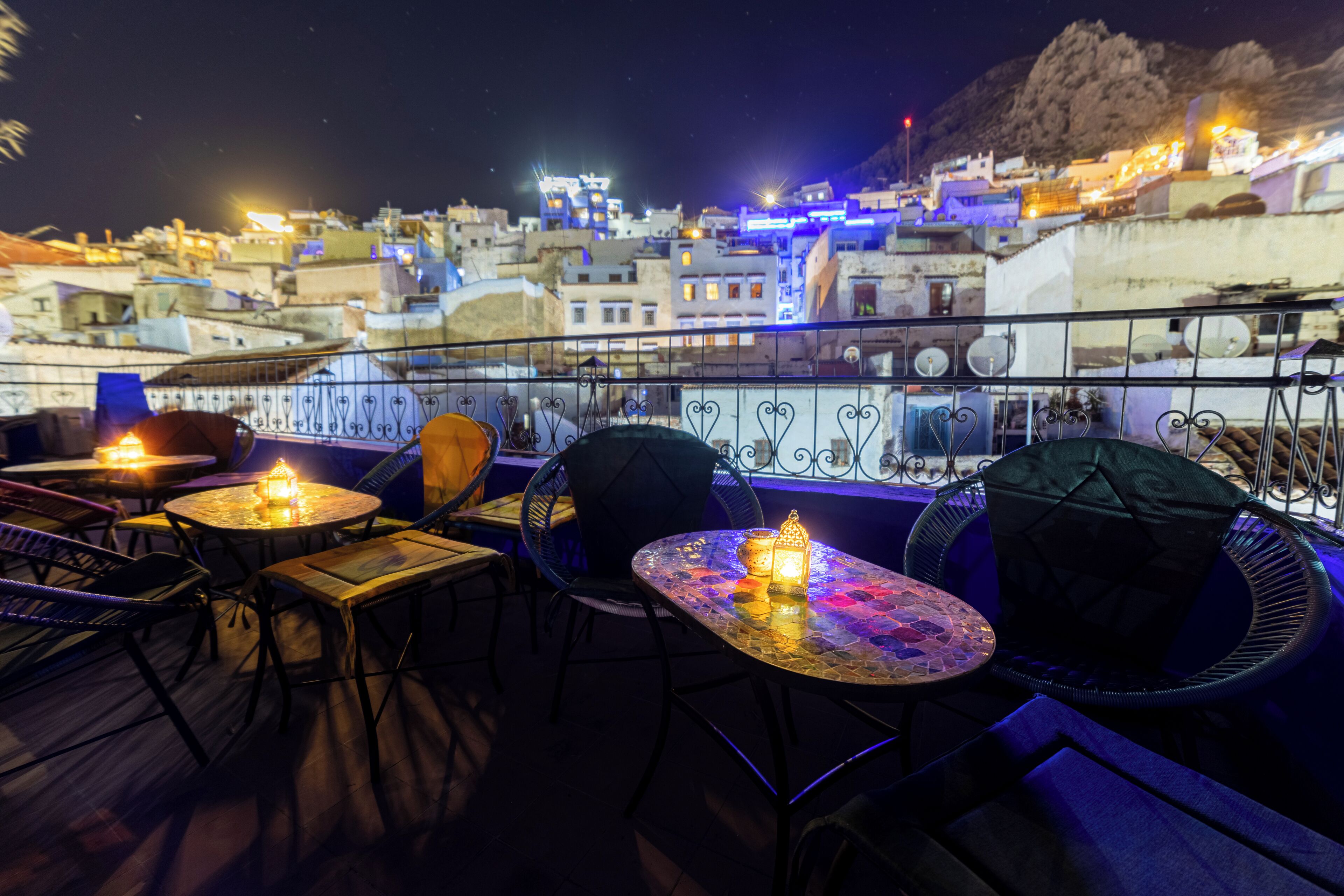 Terraza en la azotea con mesas y sillas, con vista a un paisaje urbano nocturno.