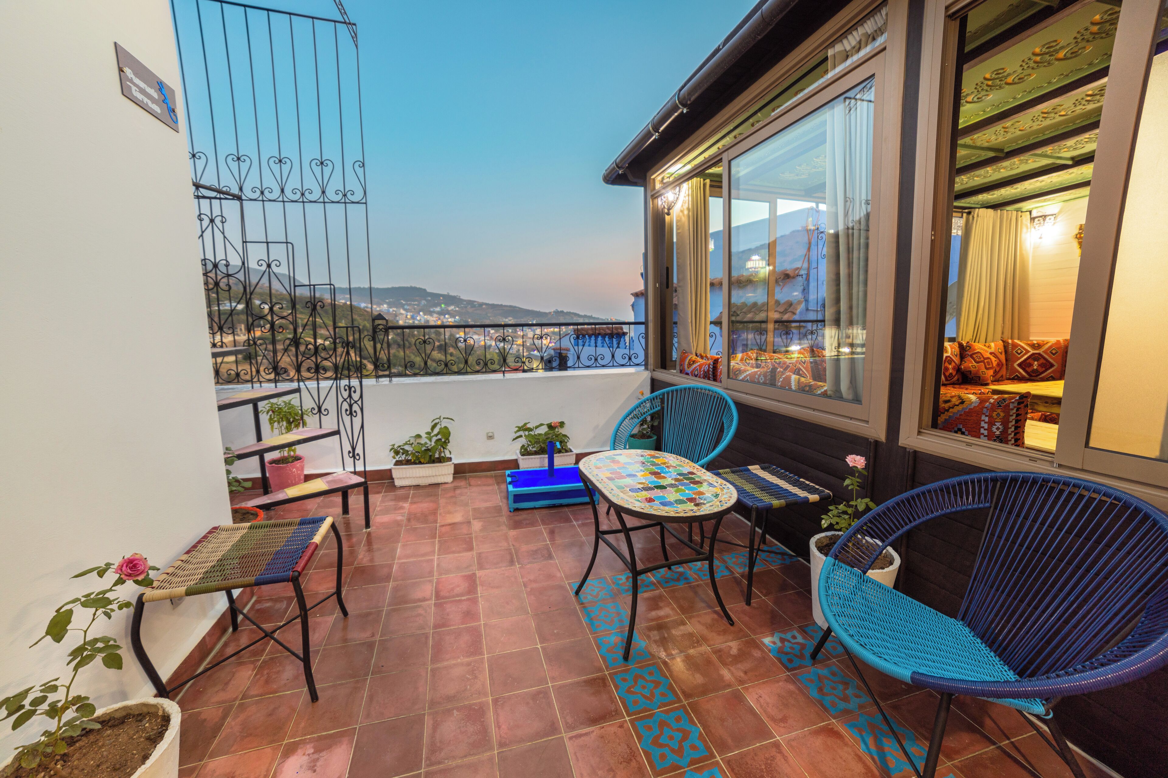A balcony with a view of the city, two blue chairs, a small table, and potted plants.