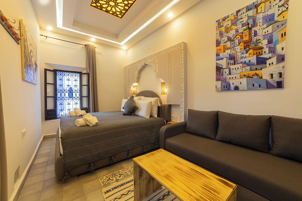 Une chambre d’hôtel moderne avec un grand lit, un canapé, une table basse en bois et un tableau représentant une ville méditerranéenne.