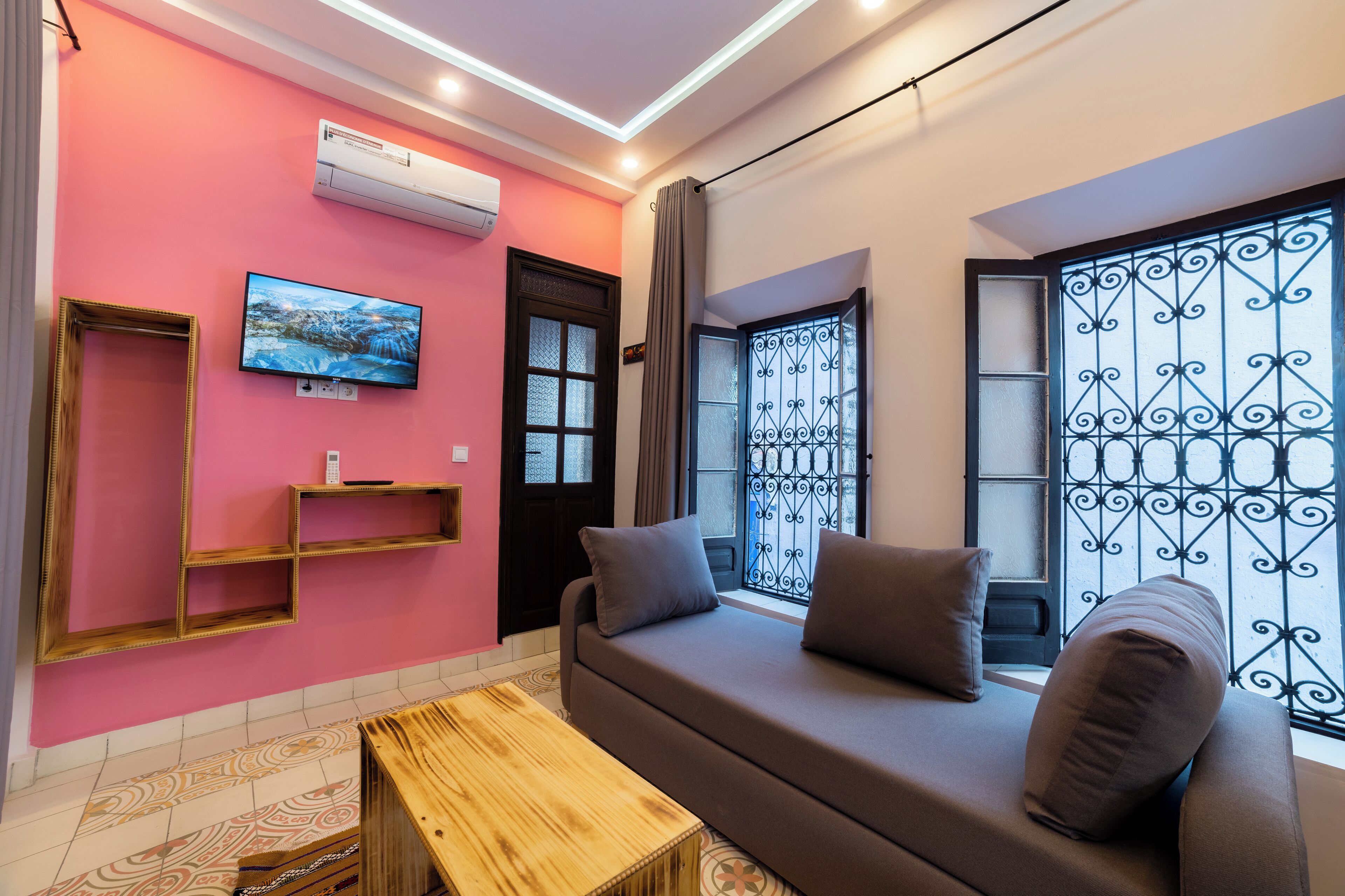 Un salón con pared rosa, sofá gris, mesa de centro de madera y televisor montado en la pared.