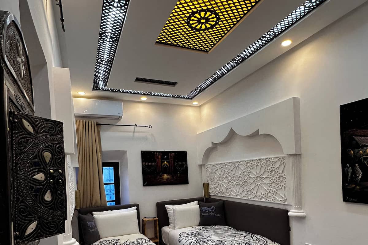 Une chambre d’hôtel moderne avec deux lits, une table en bois, des carreaux de sol à motifs, des panneaux muraux décoratifs et un plafond doté d’un éclairage sophistiqué.