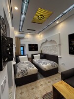 A modern hotel room with two beds, a wooden table, patterned floor tiles, decorative wall panels, and a ceiling with intricate lighting.