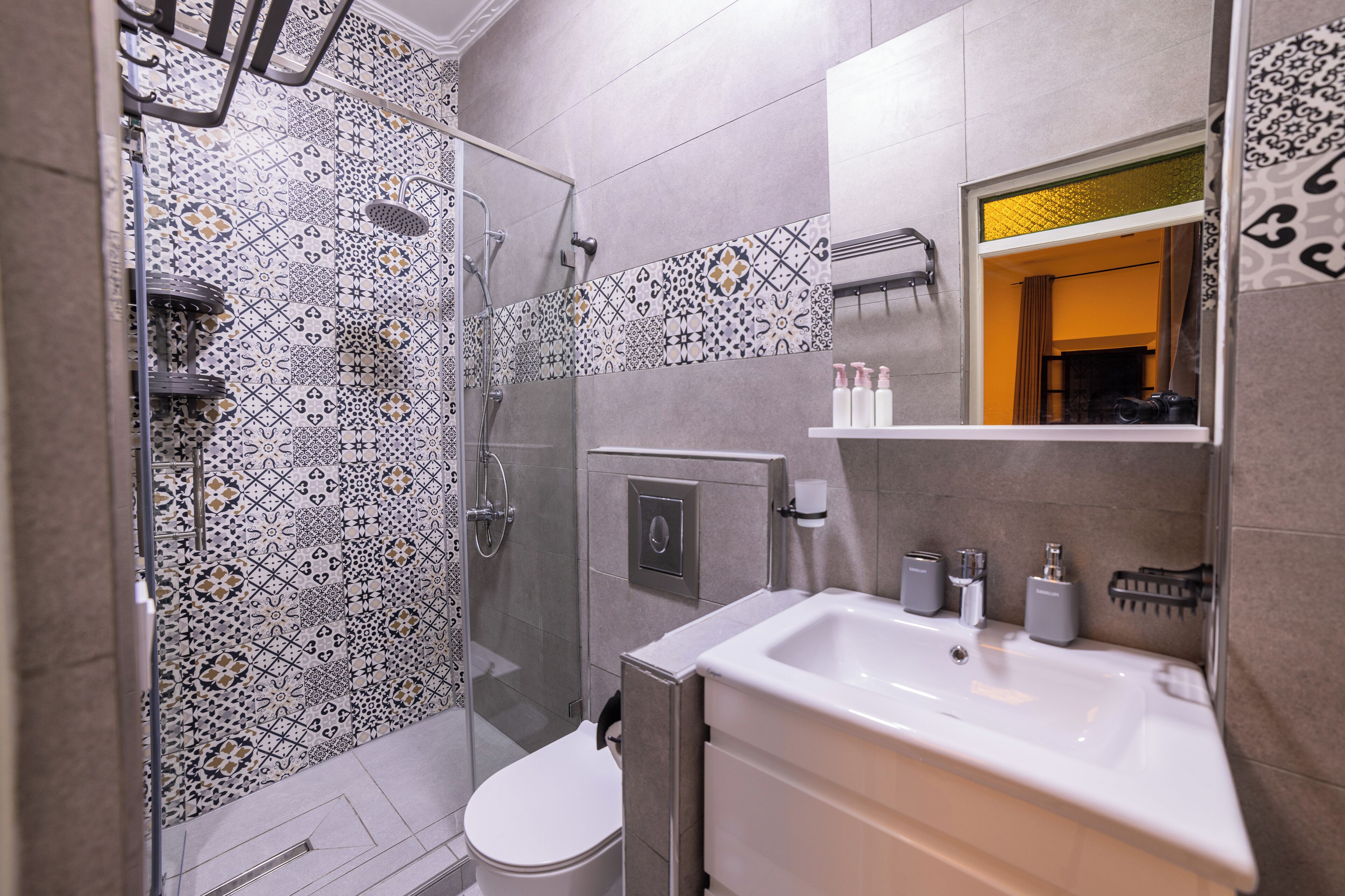 A modern bathroom with patterned tiles, a glass shower enclosure, a toilet, and a sink with toiletries.