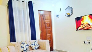 Ein Wohnzimmer mit einem beigefarbenen Sofa, einem runden Couchtisch, einem an der Wand befestigten Fernseher und einer Tür im Hintergrund.